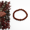 Red jasper bracelet flat in circle with deep brick-red round beads and raw jasper chunk props on light grey background