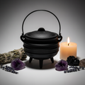 Black cast iron cauldron with lid, bail handle and three legs on light grey background with candlelight