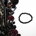 Black tourmaline icosahedron faceted beaded bracelet in circle with dark botanicals and unlit black taper on light grey background